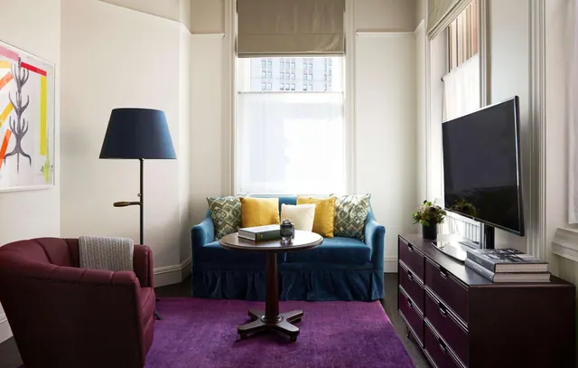 A hotel living area with a blue velvet loveseat with patterned and yellow cushions, a round wooden side table, a purple rug, a maroon armchair, a floor lamp and a flat-screen television on a dark wood dresser.