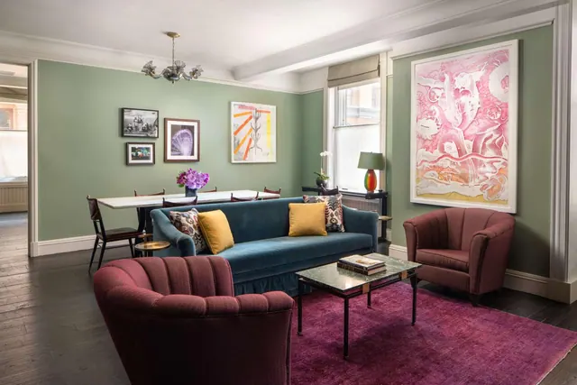 A living room with green walls, a teal velvet sofa, two maroon armchairs, a magenta rug and a large pink framed artwork, with a dining table visible in the background.