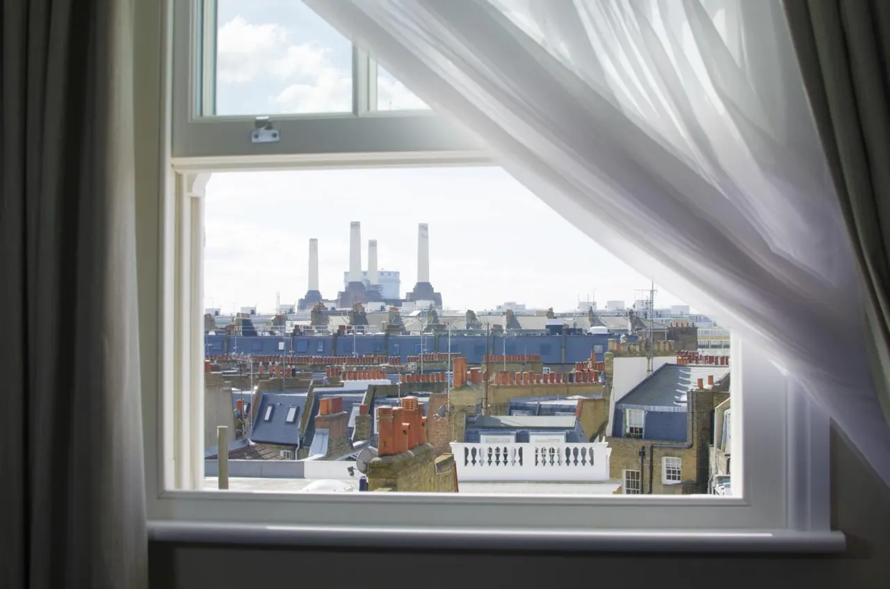 A view through a sash window over rooftops and chimneys towards Battersea Power Station, with a sheer curtain drawn across the frame.