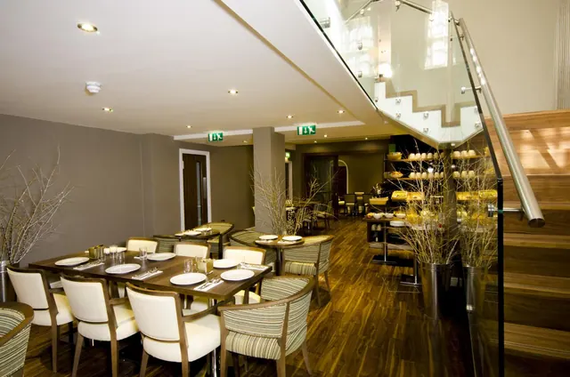 A dining room with set tables, cream upholstered chairs, a glass staircase and a buffet counter in the background.