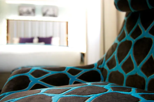 A close-up of a brown and turquoise patterned armchair in front of a blurred bed with purple cushions and framed pictures on the wall.