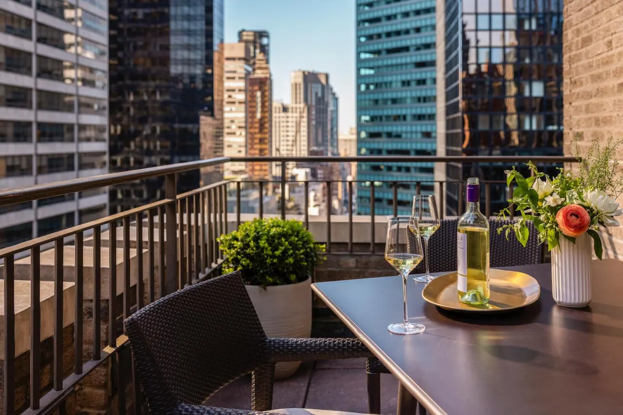 A balcony table is set with two wine glasses, a bottle of white wine on a gold tray, a flower vase and a potted shrub, with skyscrapers visible beyond the railing.