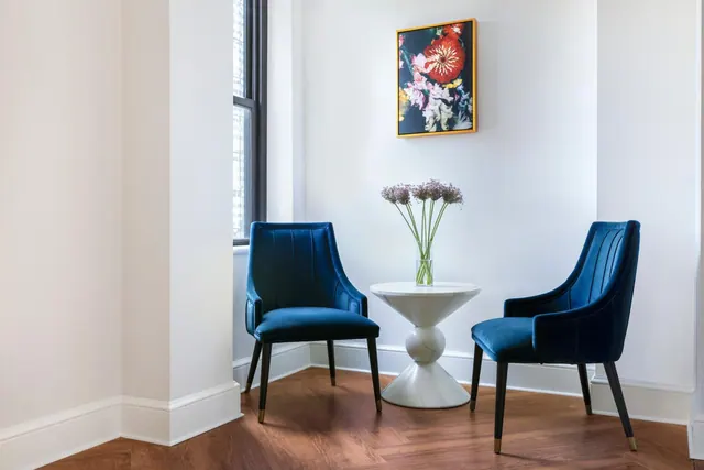 A corner seating area with two blue velvet armchairs, a white pedestal side table holding a vase of flowers, and a framed floral artwork on the wall.