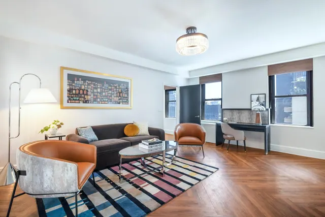 A staged living room with a sofa, two armchairs, a coffee table, a framed artwork and a desk by the windows, with neighbouring buildings visible outside.