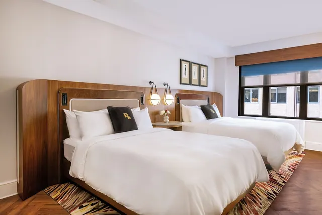 Two double beds with white linen and wooden headboards sit in a bedroom beside a window that looks onto a brick building.