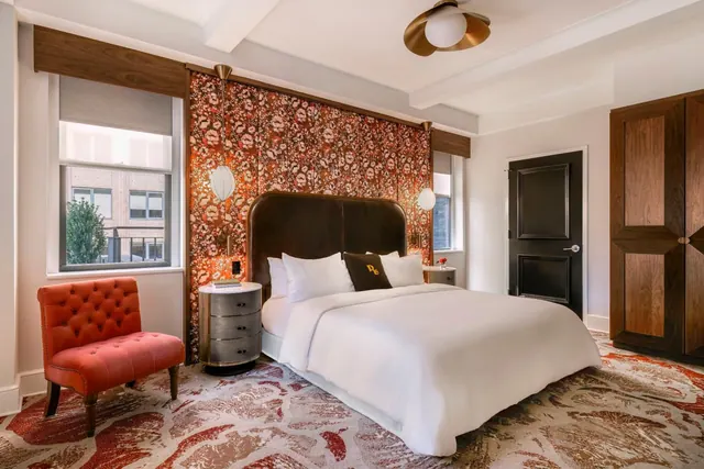 A bedroom with a king bed under white linens, a red tufted armchair, an orange floral accent wall, a wooden wardrobe and a window looking onto a brick building.