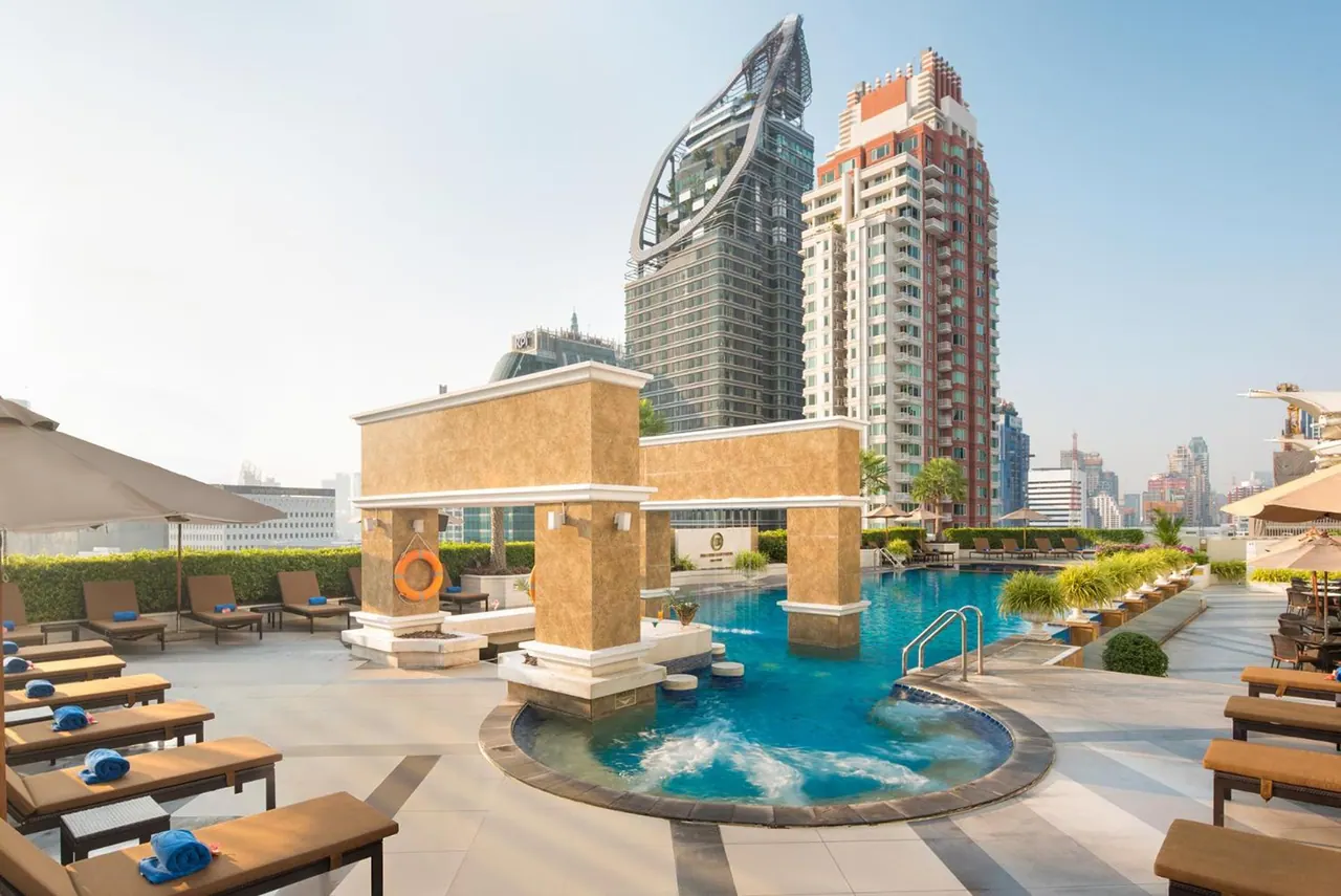 A rooftop pool with a circular bubbling spa section, sun loungers with blue towels, and tall towers in the background.