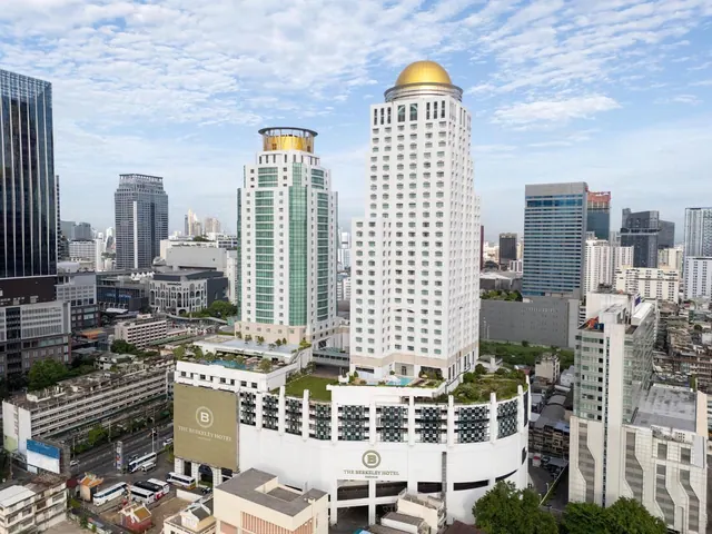Aerial view of The Berkeley Hotel's white tower with its gold dome and podium, surrounded by Bangkok high-rises under a partly cloudy sky.