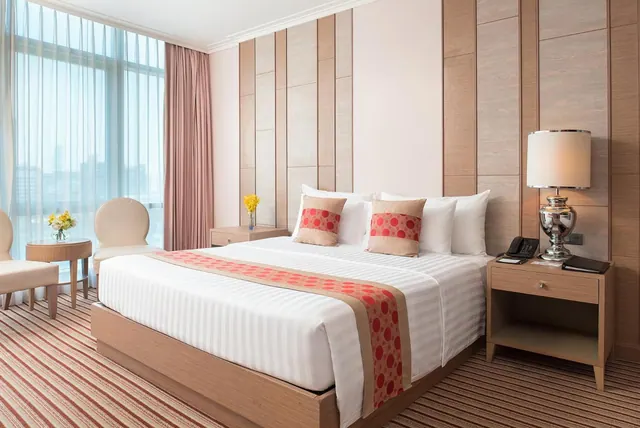 A made-up double bed with a red patterned runner sits beside a large window with sheer curtains, cream armchairs, and hazy city buildings beyond the glass.