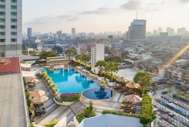 Rooftop swimming pool with a round plunge pool, sun loungers, umbrellas and outdoor dining tables, overlooking a hazy city skyline.