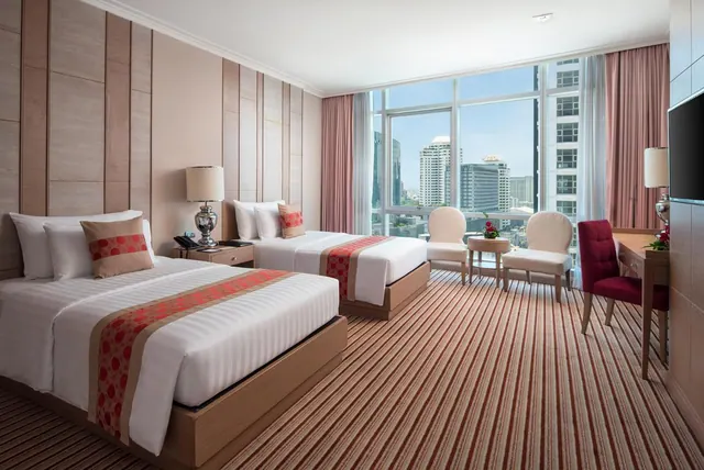 A hotel room with two beds, armchairs and a desk, with floor-to-ceiling windows overlooking high-rise buildings.