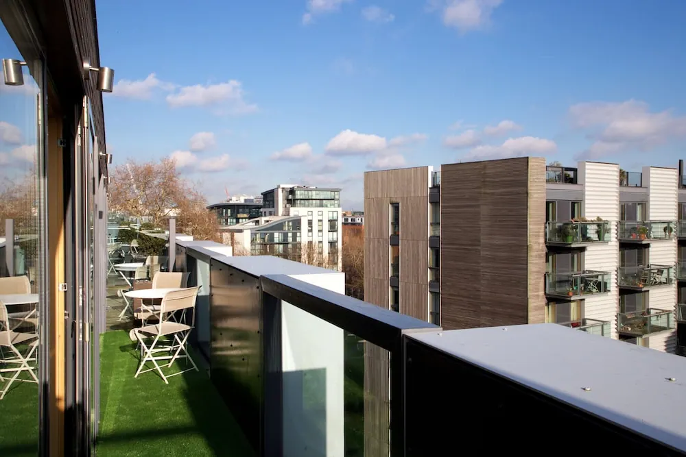 A rooftop terrace with artificial grass, folding chairs and a small table, with a glass and metal railing and apartment buildings with balconies beyond under a blue sky.