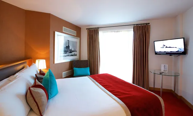 A hotel bedroom with a made double bed under a red throw, a wall-mounted television, a glass side table, a framed black-and-white print and a window with sheer curtains.