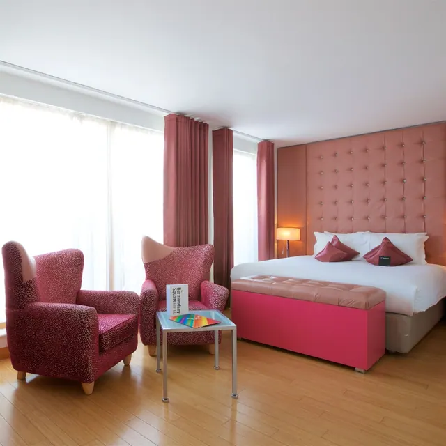A pink-themed hotel bedroom with a double bed, a tufted headboard, two leopard-print armchairs and a glass side table holding a brochure.