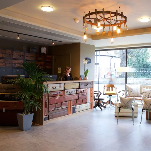 A hotel lobby with a reception desk made of stacked vintage suitcases, a woman standing behind it, a cutlery chandelier overhead, and lounge seating beside a large window onto trees.