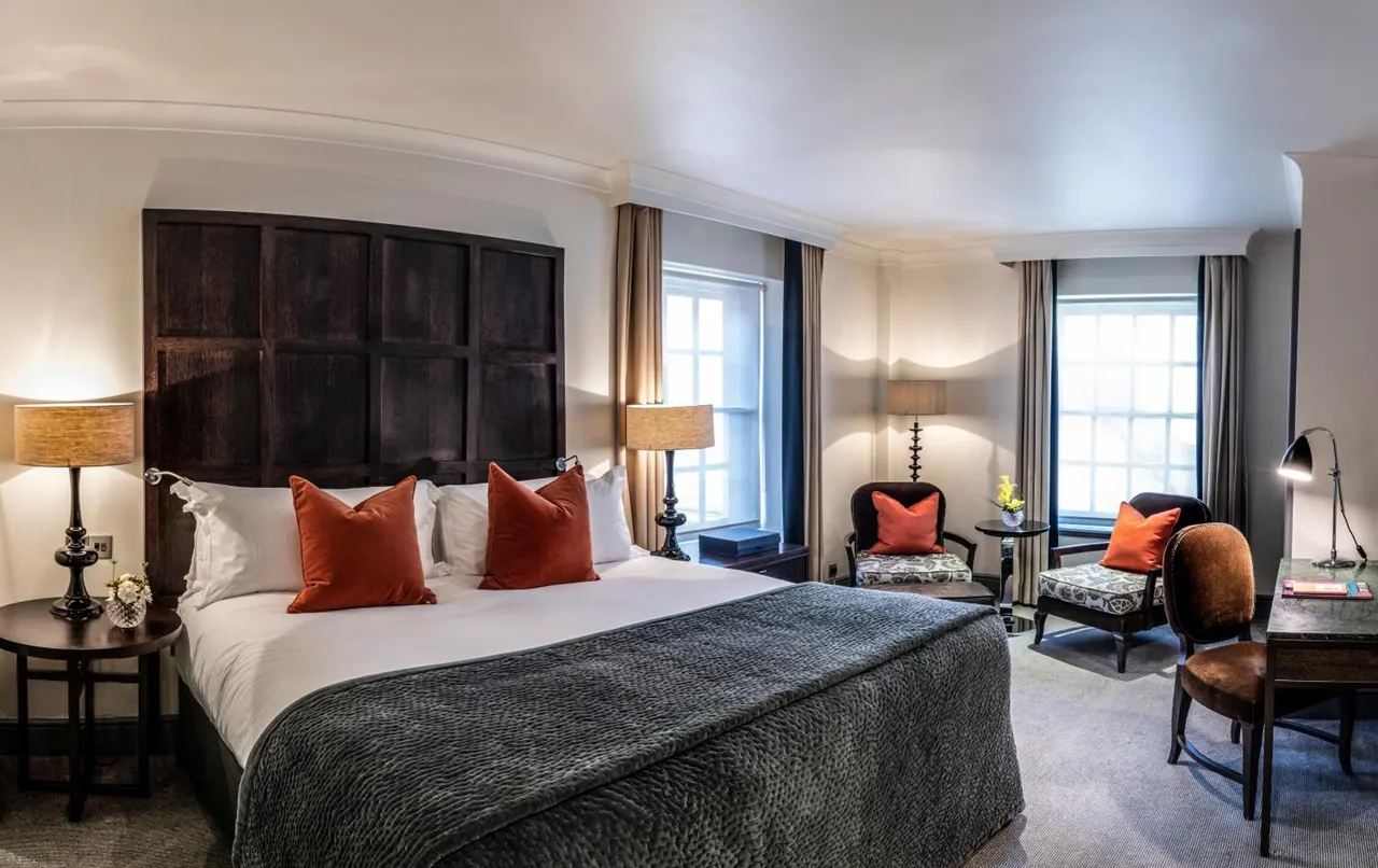 A bedroom with a large double bed with orange cushions, two armchairs by a window, and a desk on the right.