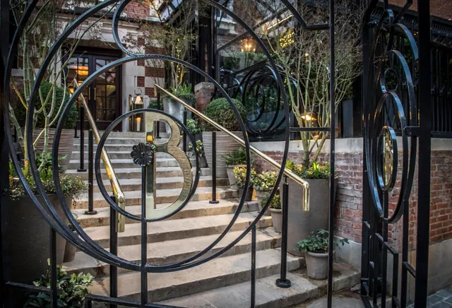An ornate black iron gate with a brass letter B stands in front of a stone staircase leading to a brick building entrance.
