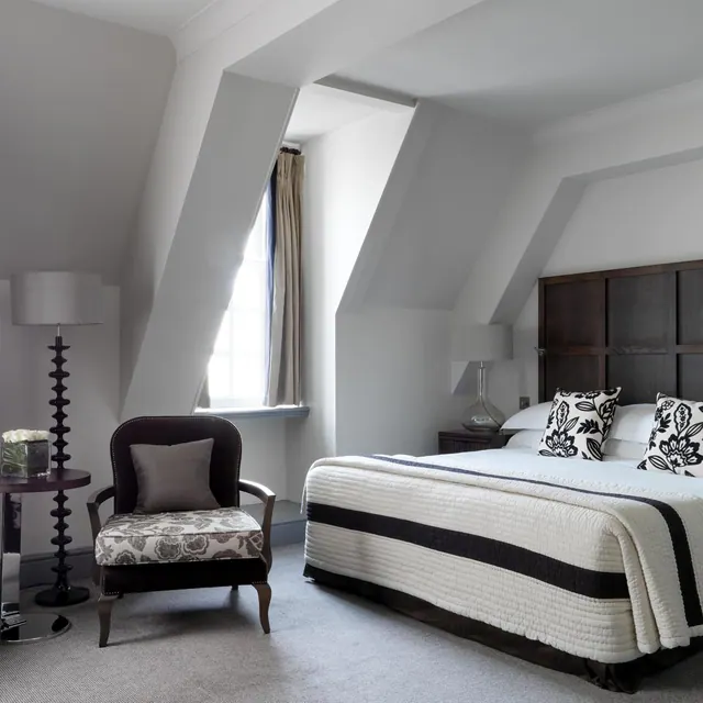 A bedroom with a made double bed, black and white cushions, an upholstered armchair, a floor lamp and a window set into a sloped ceiling.