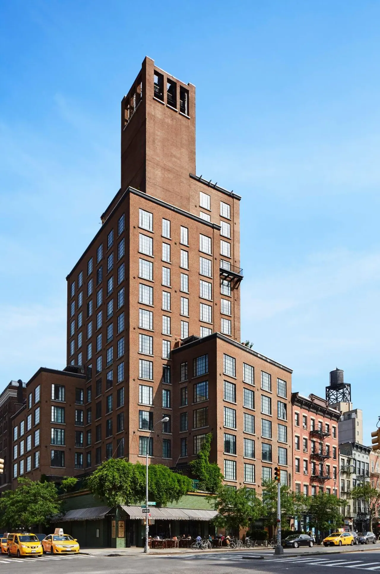 Exterior of a tall brick building with a setback tower on a street corner, with yellow taxis parked nearby and tables set out on the sidewalk under a clear blue sky.