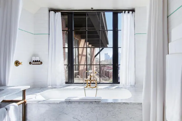 A bathroom with a marble-edged bathtub, a brass deck-mounted faucet, and a tall steel-framed window overlooking brick and city buildings.