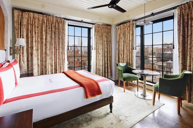 A bedroom with a made bed, two green armchairs beside a round table, a ceiling fan, and tall windows overlooking city buildings.