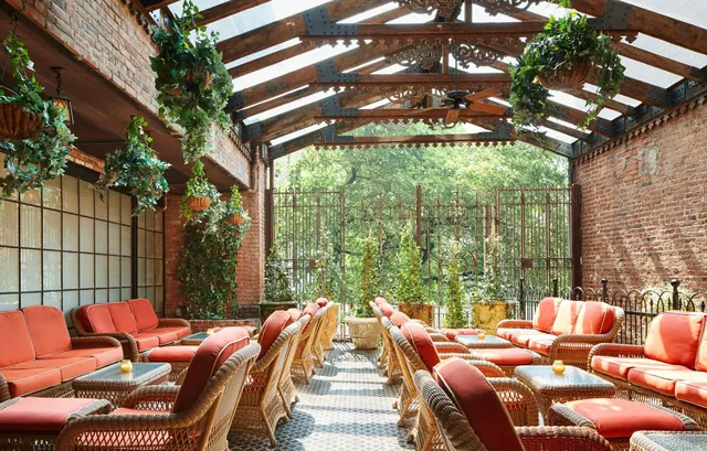 A glass-roofed brick lounge with rows of wicker chairs and red cushions, hanging plants, and an iron gate opening onto trees.
