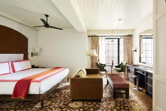 A bedroom with a king bed, a sofa and green armchairs by the window, a dark wood desk on the right, and a ceiling fan overhead.
