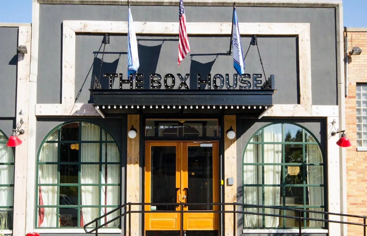 Front facade of The Box House with its name sign, three flags, arched green-framed windows and yellow double entrance doors.