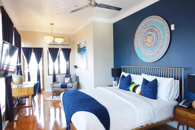 A bedroom with a king bed, a dark blue accent wall with circular wall art, a wooden desk with a globe, an armchair by the windows, and a ceiling fan.