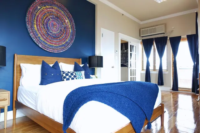 A bedroom with a wooden-framed bed dressed in white and blue linen, a dark blue accent wall with a large circular painting, blue curtains and a wall-mounted air conditioner.