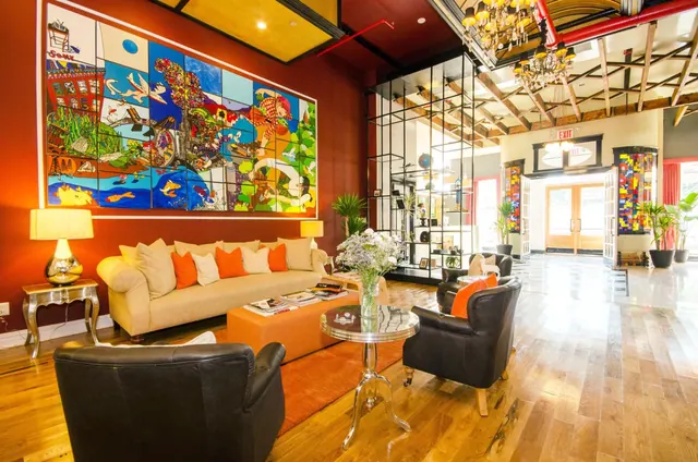 A lobby seating area with a red wall, a large colorful mural, a beige sofa, leather armchairs and a round glass side table, with chandeliers hanging beneath an exposed timber ceiling.