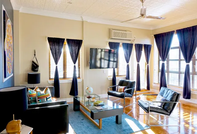 A living room with a black leather sofa, two leather armchairs, a glass coffee table on a blue rug, a wall-mounted television and navy curtains at tall windows.