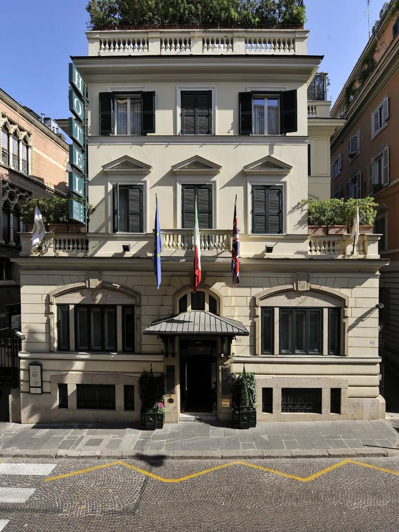 The front facade of a cream-coloured hotel building with dark green shutters, a canopy over the entrance, flags on the upper floor and a vertical HOTEL sign on the left, under a clear blue sky.
