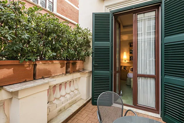 A balcony with terracotta planters, a stone balustrade and a metal chair and table, with open glass doors leading into a bedroom with a lamp and bed.