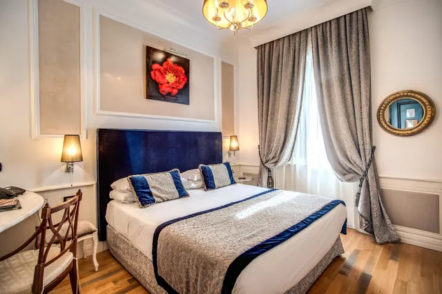 A bedroom with a double bed under a navy velvet headboard, flanked by wall lamps, with a desk and chair on the left and a framed red flower painting above the bed.