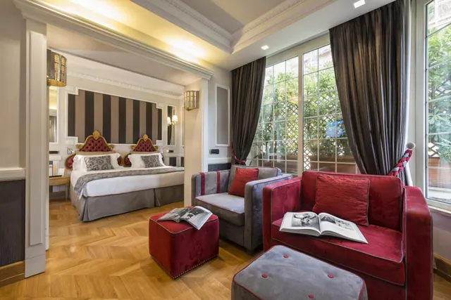 A staged suite with red and grey armchairs and ottomans in the foreground, a bed visible through an archway, and tall windows looking onto green plants.