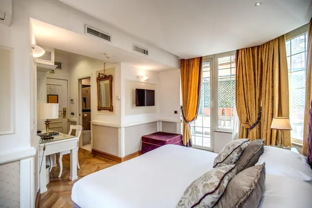 A bedroom with a white double bed, gold curtains, a wall-mounted television, a writing desk and an open doorway to a bathroom.
