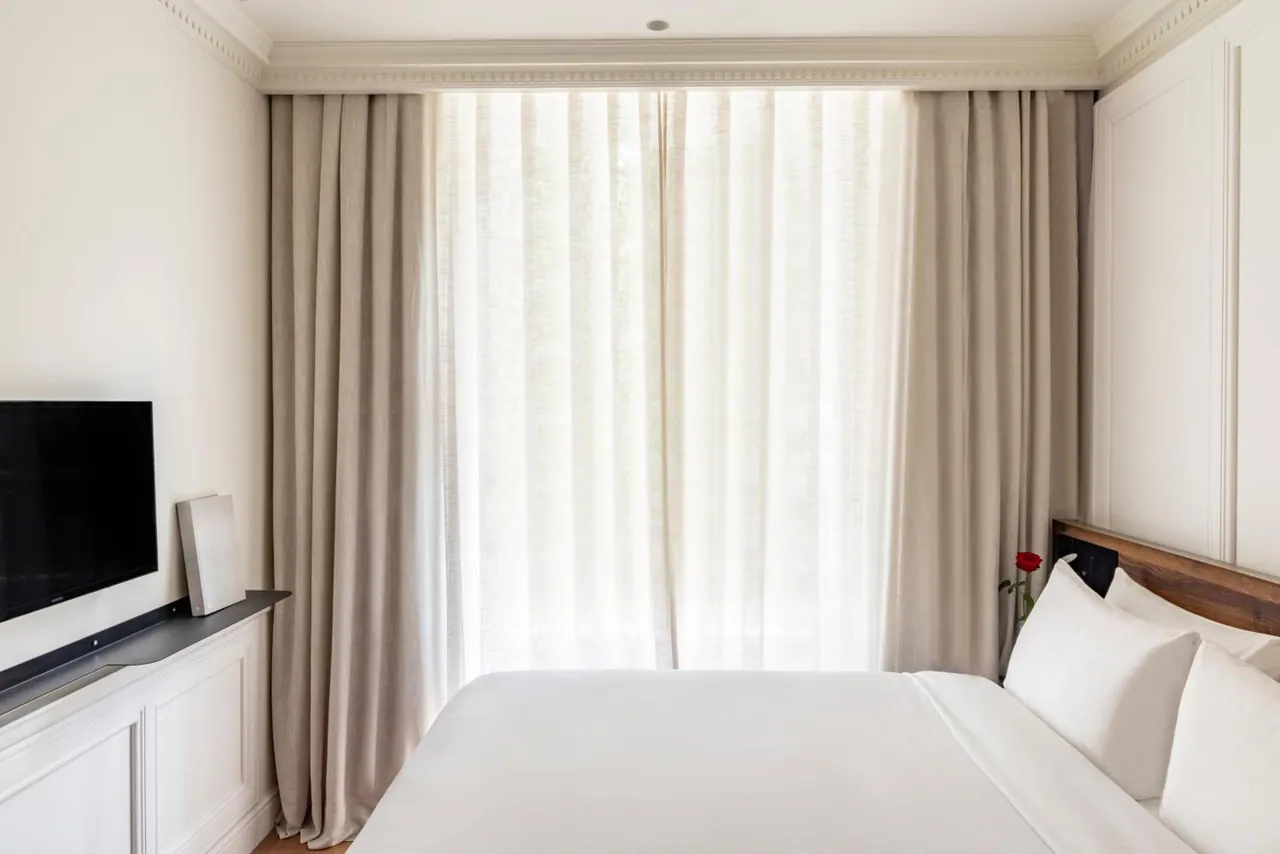 A bedroom with a made white bed, sheer and linen curtains over a window, and a flat-screen television on a white cabinet.
