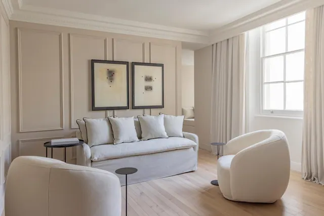 A living room with a light grey sofa, two white curved armchairs, small black side tables, framed artwork on a panelled wall and a light wood floor.