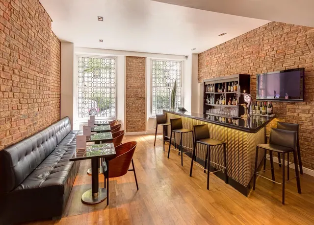A bar area with exposed brick walls, a tufted leather banquette, high bar stools, a wall-mounted television and tall windows with patterned glass.