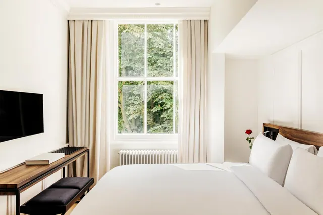 A bedroom with a made double bed, a wall-mounted television, a wooden desk with a book on it, and a window looking onto green trees.