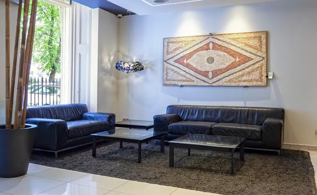 A lobby seating area with two black leather sofas, low glass coffee tables, a mosaic wall artwork and a tall window showing trees and railings outside.