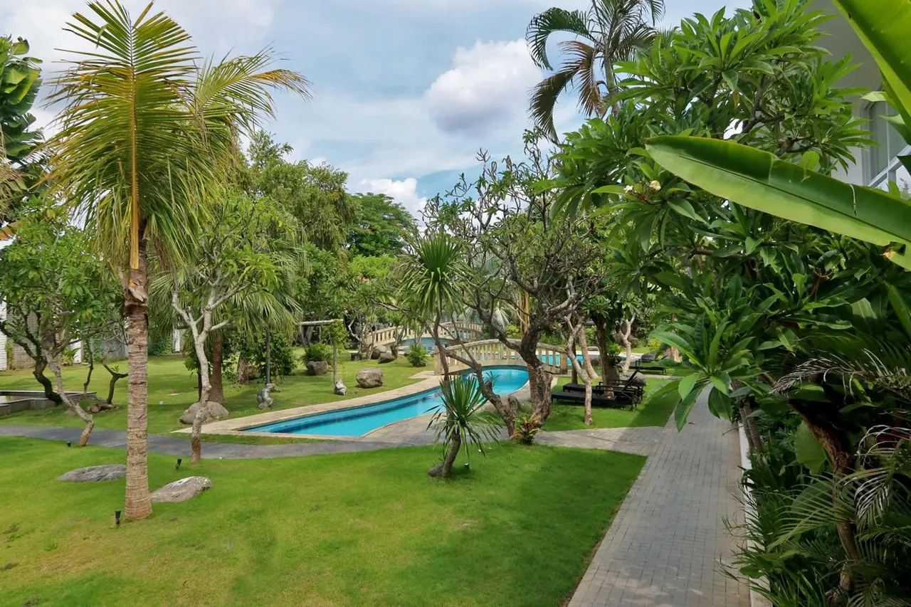 A kidney-shaped swimming pool sits among palm and frangipani trees and a trimmed lawn, with dark sun loungers beside it and a brick path running along the right edge.