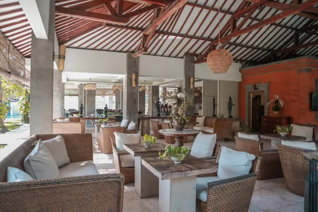 An open-air lounge with rattan armchairs and sofas with white cushions, wooden tables, and a red-beamed timber roof.