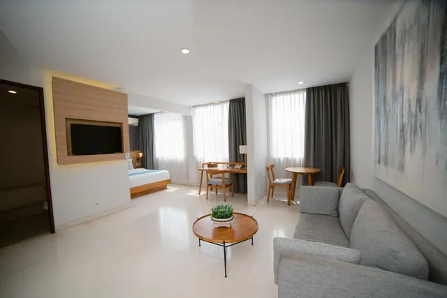 A living area with a grey sofa, a round wooden coffee table, a wall-mounted television, and a bed and writing desk visible in the adjoining space.