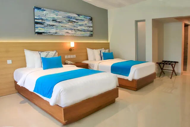 A bedroom with two beds dressed in white linen with blue runners, a large abstract painting on the wall, and a luggage stand near the doorway.