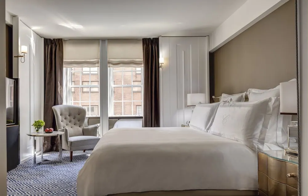 A bedroom with a king bed, a grey armchair by the window, a small side table with flowers, and a television on the left wall, with a view of a brick building through the windows.