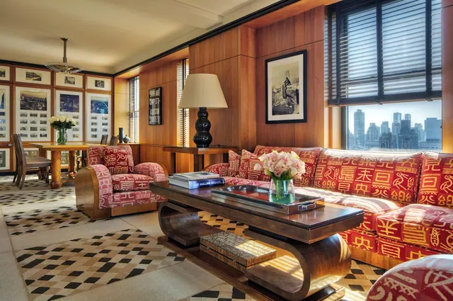 A living room with a red patterned sofa and armchairs, a wooden coffee table with a flower vase, a table lamp, wood-panelled walls and a window showing a city skyline.
