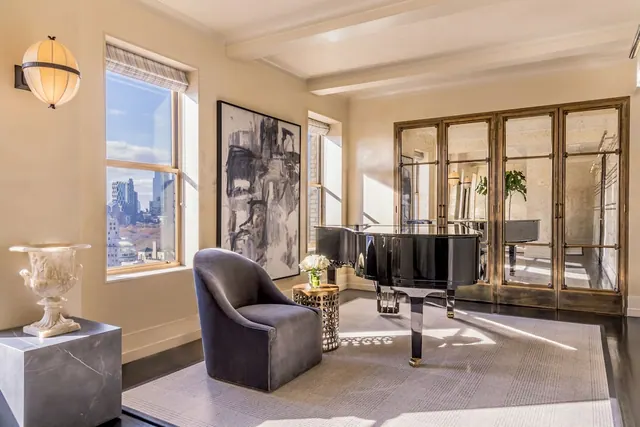 A sunlit living room with a black grand piano, a grey armchair, a large abstract painting, and a window looking out over city buildings.