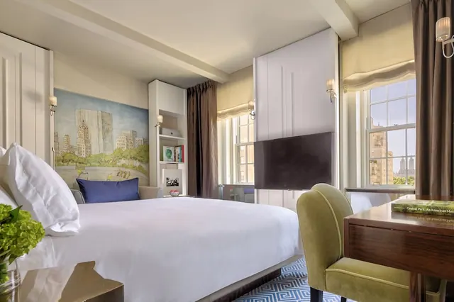 A bedroom with a made king bed, a wall-mounted television, a painted city mural, and a green chair at a wooden desk by a window overlooking buildings.